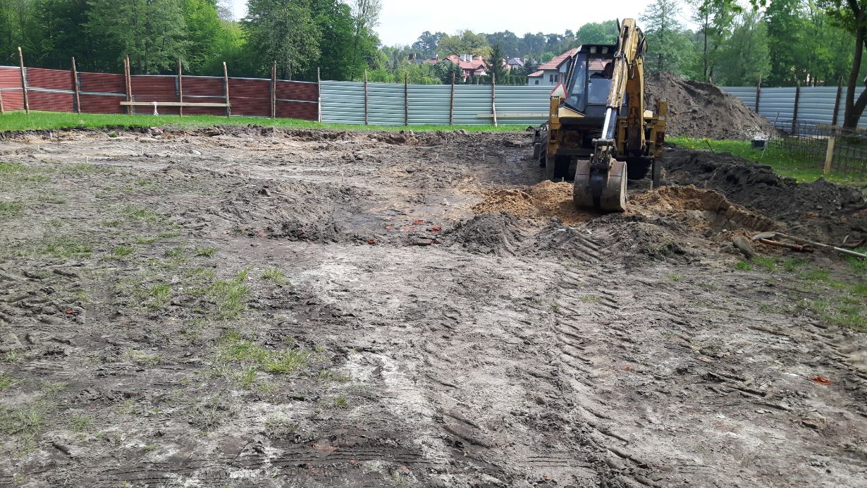 Zdjęcie przedstawia plac budowy z koparką w trakcie prac ziemnych. W tle widać ogrodzenie z blachy falistej oraz drzewa. Na pierwszym planie widoczne są ślady po ciężkim sprzęcie na ziemi.