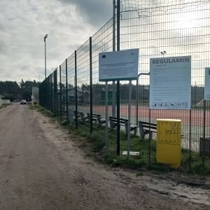 Zdjęcie przedstawia ogrodzone boisko sportowe z tablicami informacyjnymi. Na pierwszym planie widoczna jest żółta skrzynka oraz tablica z napisem "REGULAMIN". W tle widać ławki, boisko oraz latarnie.