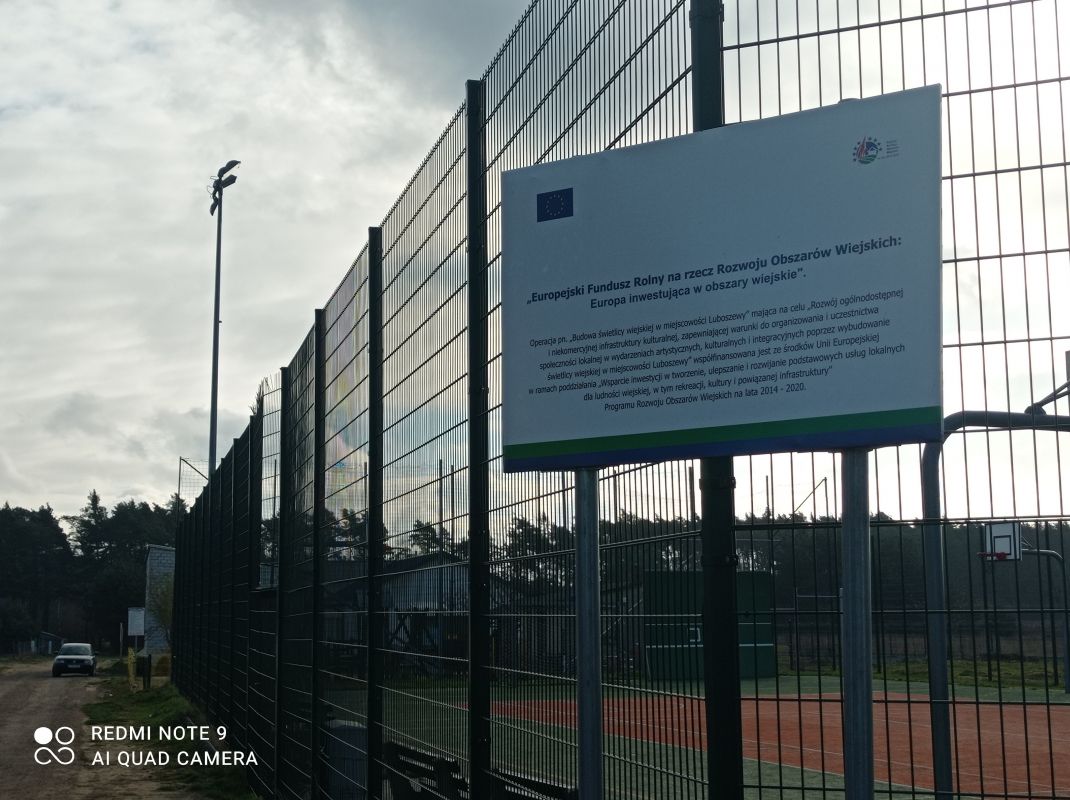 Zdjęcie przedstawia ogrodzenie boiska sportowego z zamontowaną tablicą informacyjną. Na tablicy znajduje się tekst dotyczący Europejskiego Funduszu Rolnego na rzecz Rozwoju Obszarów Wiejskich, który inwestuje w obszary wiejskie. W tle widać latarnię i drzewa.