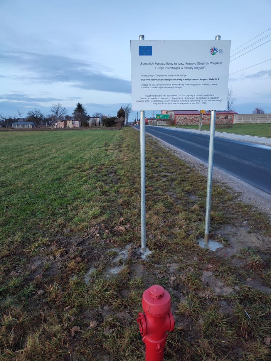 Zdjęcie przedstawia znak informacyjny na tle wiejskiego krajobrazu. Na pierwszym planie widoczny jest czerwony hydrant przeciwpożarowy. Znak informacyjny jest umieszczony na dwóch metalowych słupkach i zawiera tekst w języku polskim oraz logo Unii Europejskiej i Programu Rozwoju Obszarów Wiejskich. W tle widać drogę asfaltową, trawnik oraz kilka budynków i drzew.