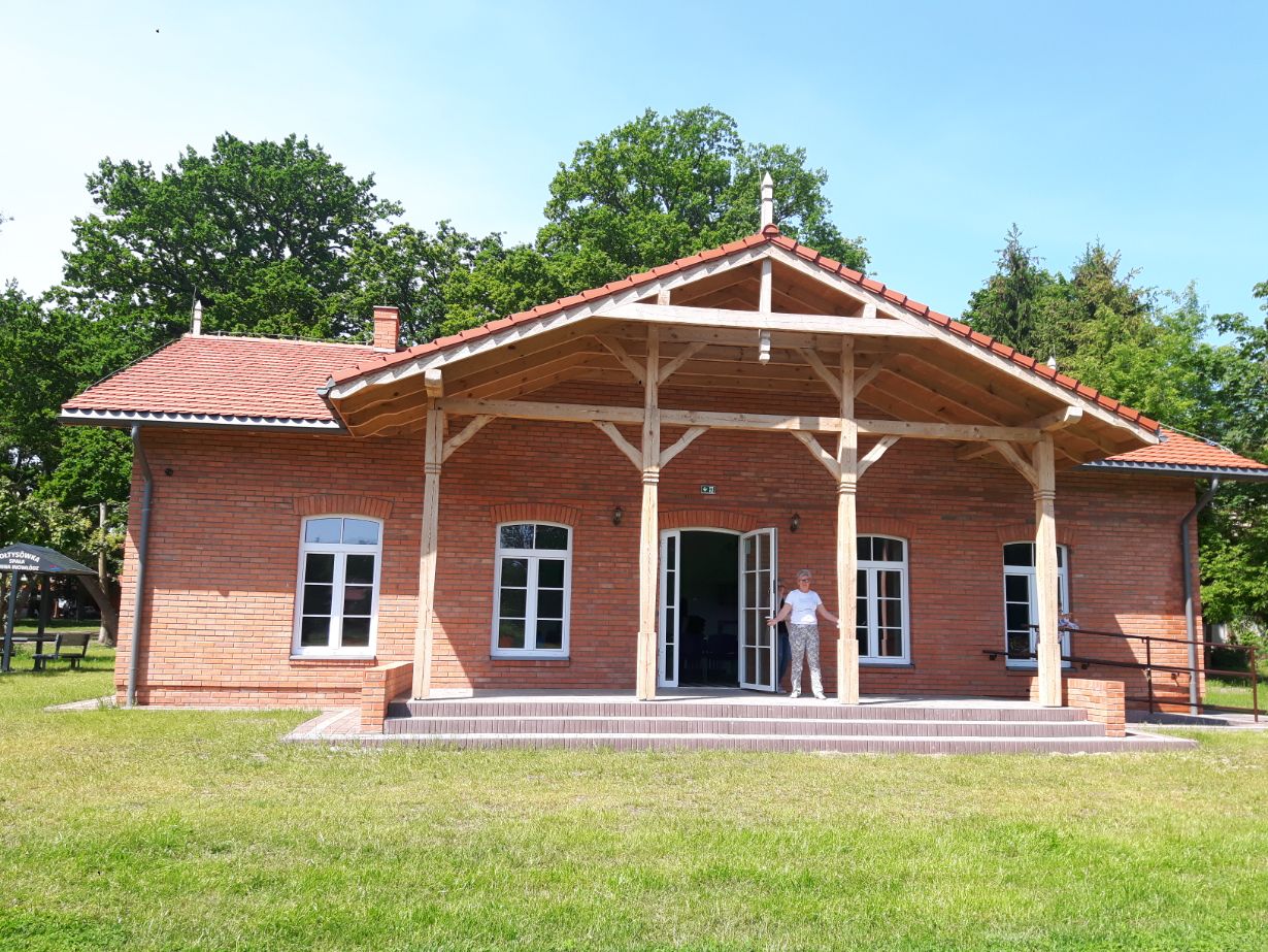 Zdjęcie przedstawia ceglaną budowlę z drewnianym zadaszeniem nad wejściem. Przed budynkiem stoi osoba w białej koszulce i jasnych spodniach. W tle widać drzewa i trawnik.