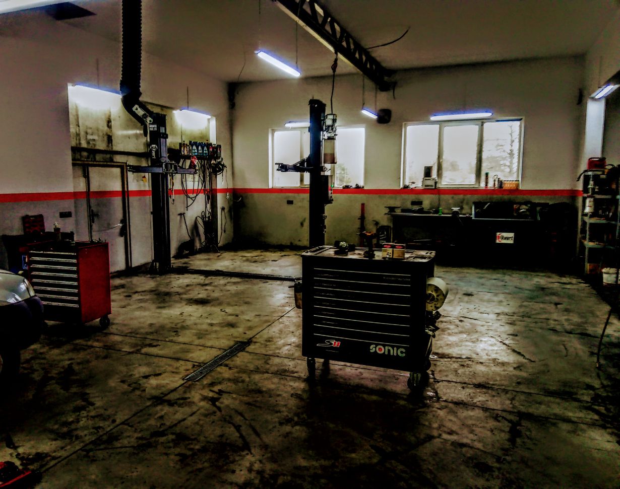 Zdjęcie przedstawia warsztat samochodowy z podnośnikiem hydraulicznym, narzędziami i sprzętem. Na środku znajduje się wózek narzędziowy marki Sonic. W tle widać okna, półki z narzędziami i różne urządzenia. Pomieszczenie jest dobrze oświetlone lampami sufitowymi.
