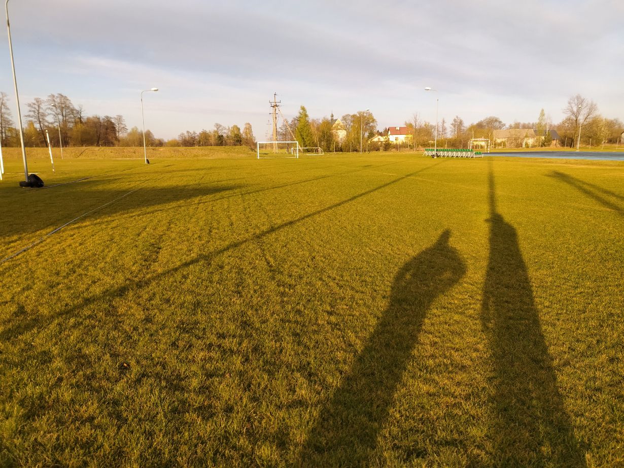 Na zdjęciu widać boisko sportowe, prawdopodobnie do piłki nożnej, z widocznymi bramkami w tle. Wokół boiska znajdują się wysokie latarnie, a na trawie rzucają się w oczy cienie dwóch osób, co sugeruje, że zdjęcie zostało zrobione późnym popołudniem lub wczesnym rankiem, gdy słońce jest nisko. W tle widać drzewa, kilka budynków oraz niebieską bieżnię lub pole.