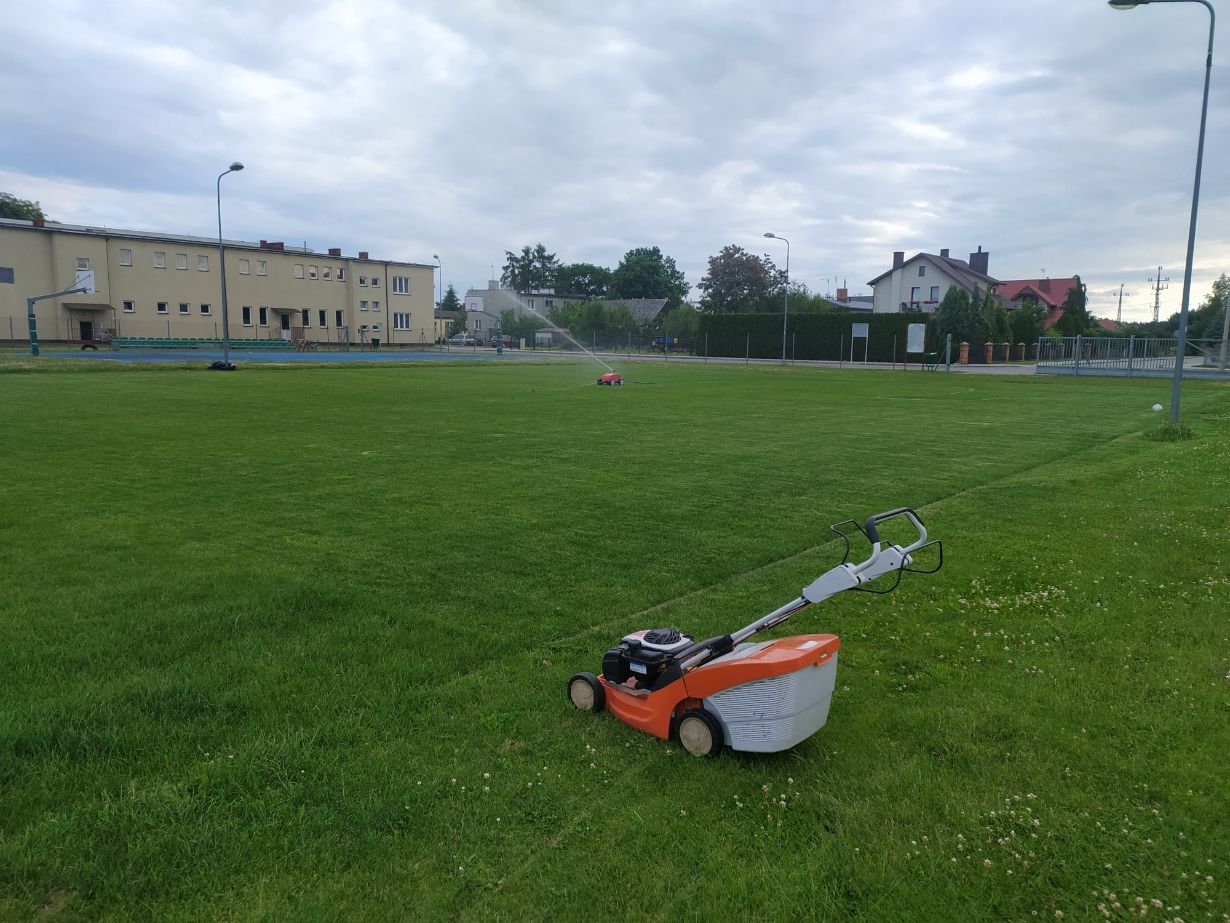 Na zdjęciu widać kosiarkę do trawy stojącą na dużym, zielonym trawniku. W tle znajduje się budynek, kilka latarni ulicznych oraz inne domy.