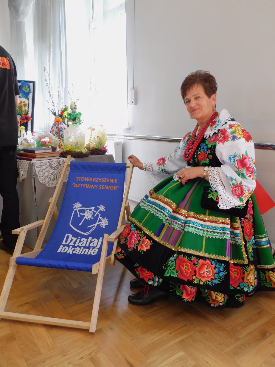 Kobieta w kolorowym stroju ludowym siedzi na krześle z napisem "STOWARZYSZENIE AKTYWNY SENIOR Działaj lokalnie". Obok znajduje się stół z ozdobami wielkanocnymi.