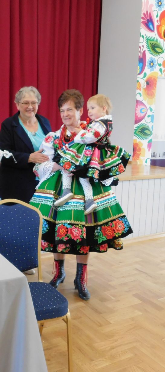 Trzy osoby, w tym dziecko, ubrane w tradycyjne stroje ludowe, stoją na tle czerwonej zasłony i kolorowej ściany. Stroje są bogato zdobione kwiatowymi wzorami i koralikami.