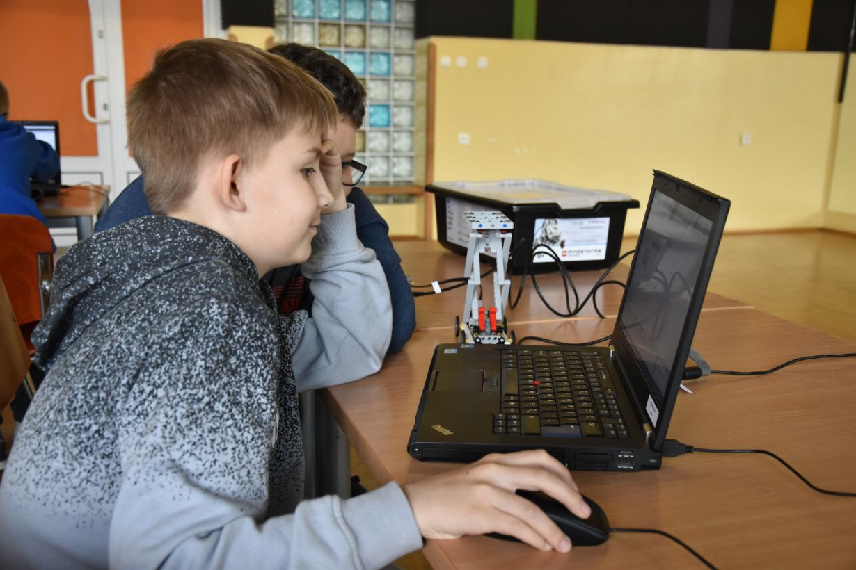 Na zdjęciu widać dwóch chłopców siedzących przy biurku, patrzących na ekran laptopa. Jeden z nich trzyma myszkę komputerową. Na biurku znajduje się mały robot zbudowany z klocków LEGO, podłączony do laptopa. W tle widać inne dzieci i sprzęt komputerowy.