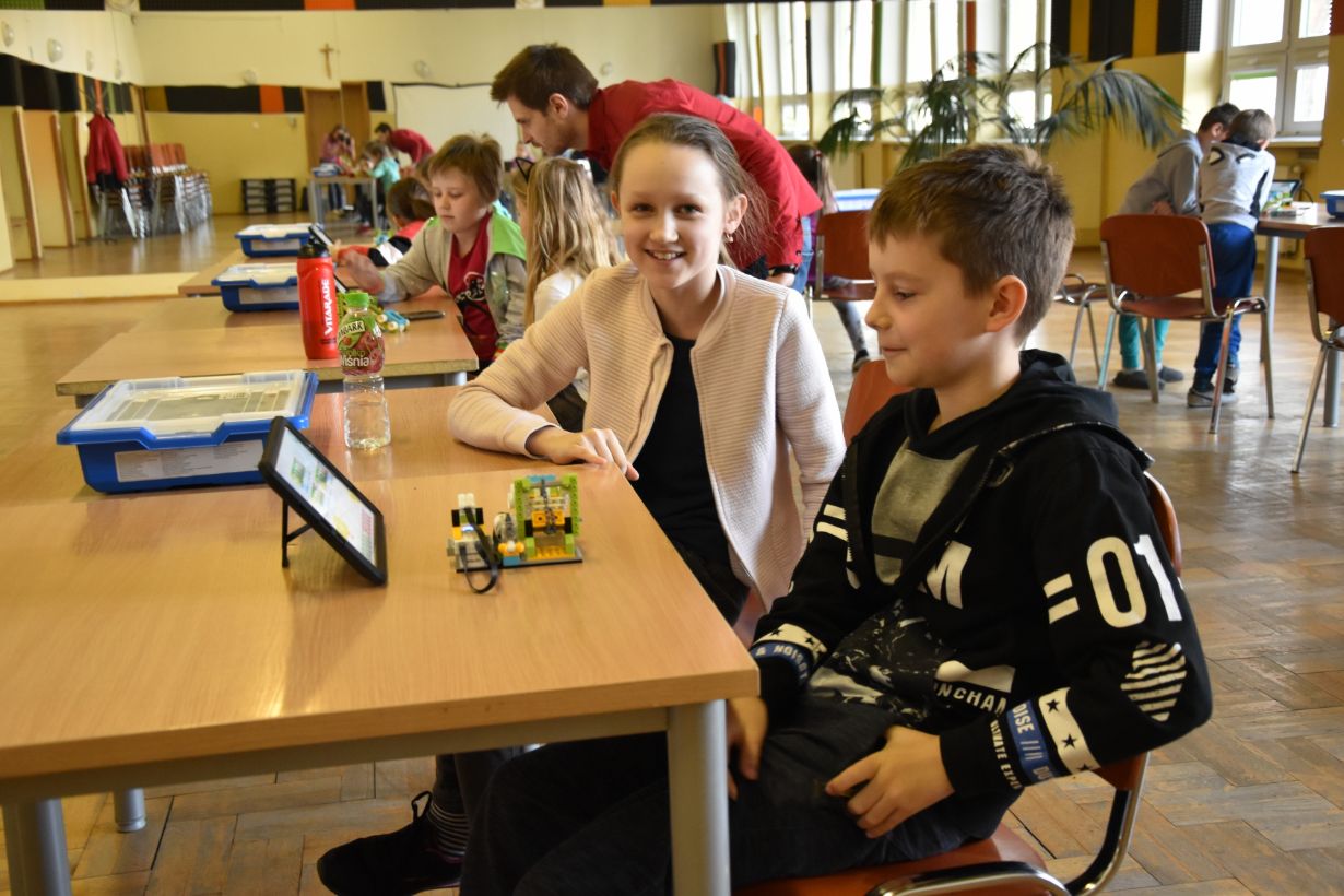 W sali lekcyjnej dzieci siedzą przy stołach, pracując nad projektami z klocków LEGO i tabletami. Na pierwszym planie dwójka dzieci siedzi przy stole z tabletem i konstrukcją LEGO. W tle widać inne dzieci i nauczyciela pomagającego uczniom.