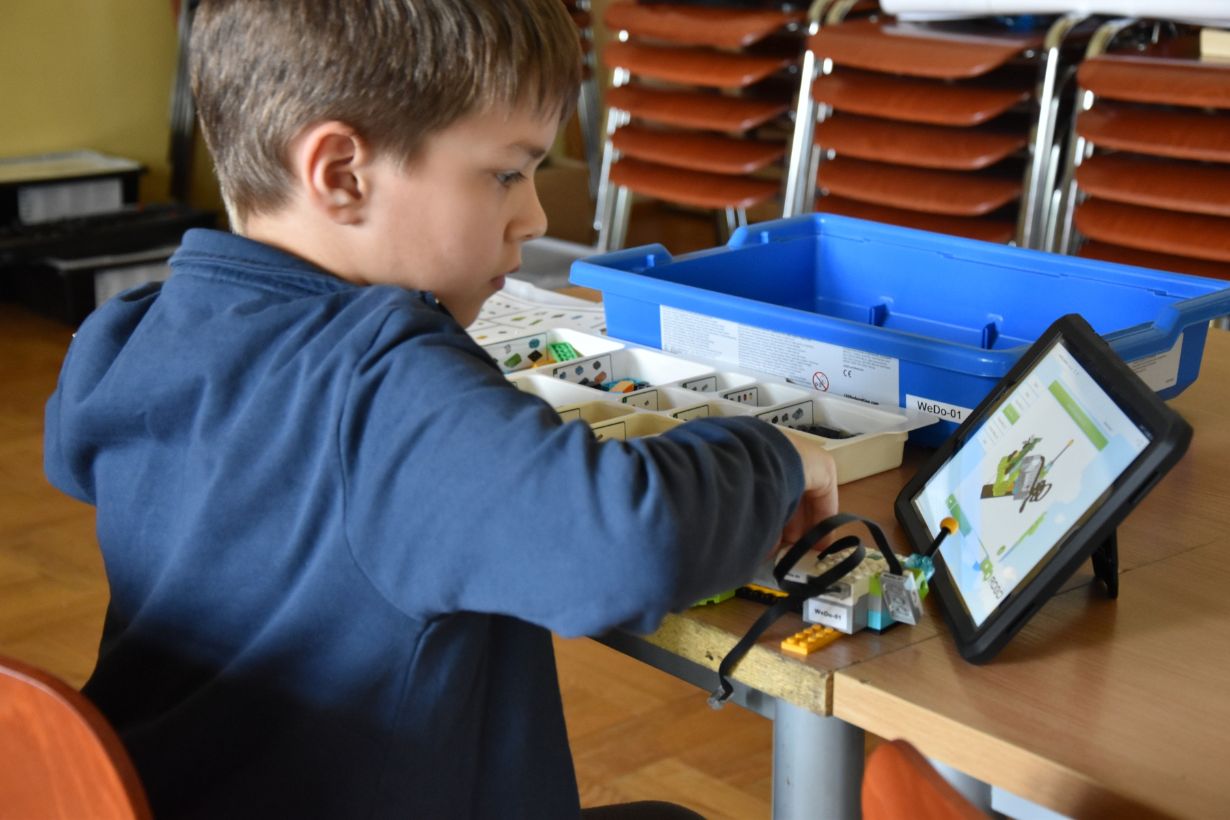 Dziecko siedzi przy stole, korzystając z tabletu i zestawu klocków edukacyjnych WeDo 2.0. Na ekranie tabletu widoczna jest instrukcja budowy modelu z klocków. Na stole znajduje się niebieskie pudełko z przegródkami na klocki. W tle widać rząd krzeseł.