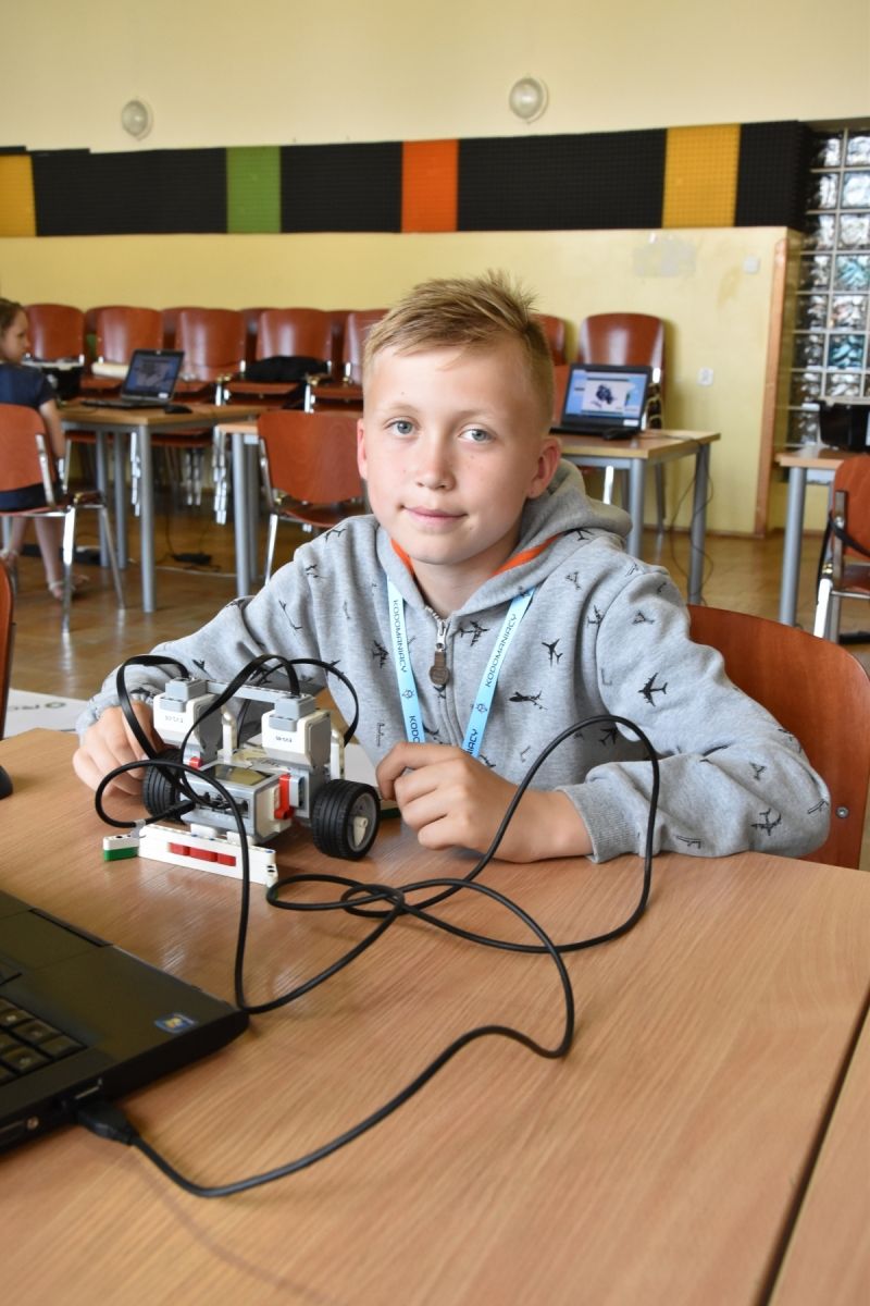Na zdjęciu widać chłopczyka siedzącego przy stole w sali lekcyjnej, trzymającą robota zbudowanego z klocków LEGO Mindstorms. Na stole znajduje się laptop, do którego robot jest podłączony za pomocą kabli. W tle widać inne stoły, krzesła i laptopy.