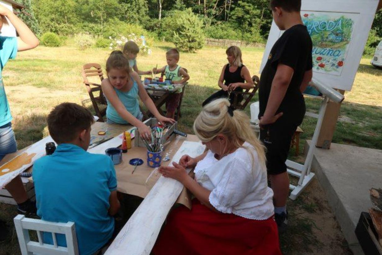 Obraz przedstawia grupę osób siedzących przy stole na świeżym powietrzu, zajmujących się malowaniem i rysowaniem. Na stole znajdują się farby, pędzle i inne przybory plastyczne. Obrazek przedstawia kreatywne zajęcia plastyczne na świeżym powietrzu.