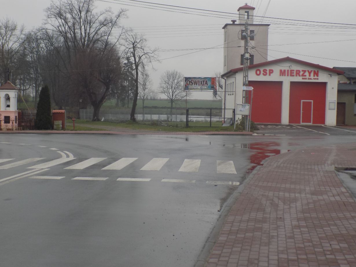 Zdjęcie przedstawia skrzyżowanie z przejściem dla pieszych, na którym widać budynek straży pożarnej OSP Mierzyn z czerwonymi drzwiami garażowymi i wieżą. W tle znajduje się billboard z napisem "OŚWIATA" oraz drzewa i pole. Po lewej stronie widoczna jest mała kapliczka.