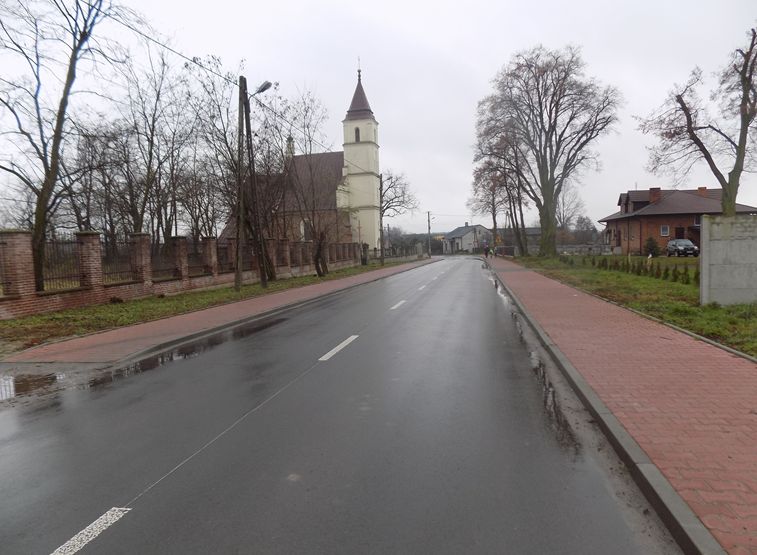 Zdjęcie przedstawia pustą, mokrą ulicę z chodnikami po obu stronach. Po lewej stronie znajduje się kościół z wieżą, otoczony murem z cegły. Po prawej stronie widoczny jest dom z czerwonej cegły oraz drzewa bez liści.