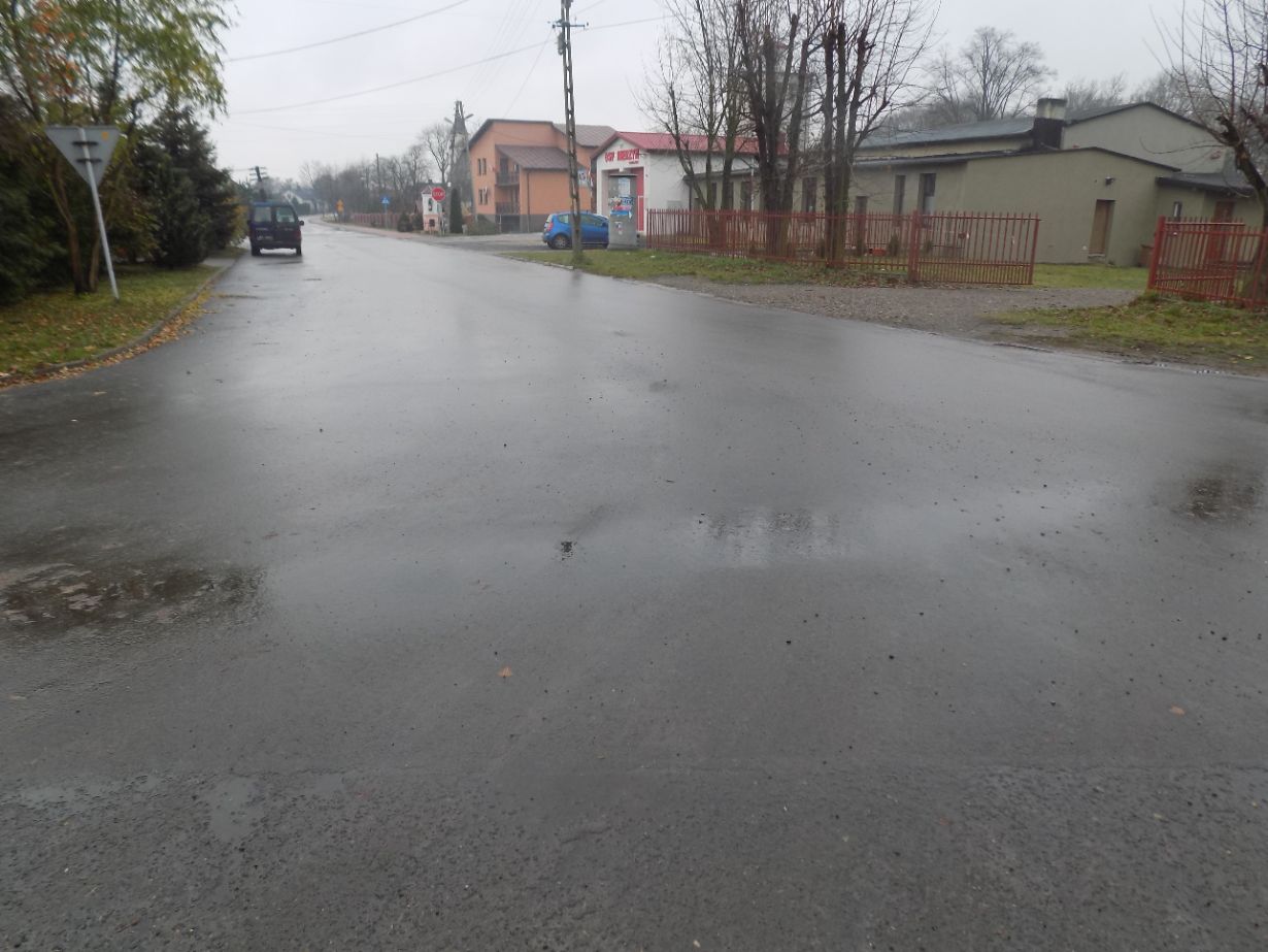 Zdjęcie przedstawia mokrą, asfaltową drogę na skrzyżowaniu w małej miejscowości. Po lewej stronie znajduje się znak drogowy, a w tle widać zaparkowane samochody, budynki mieszkalne oraz drzewa bez liści.