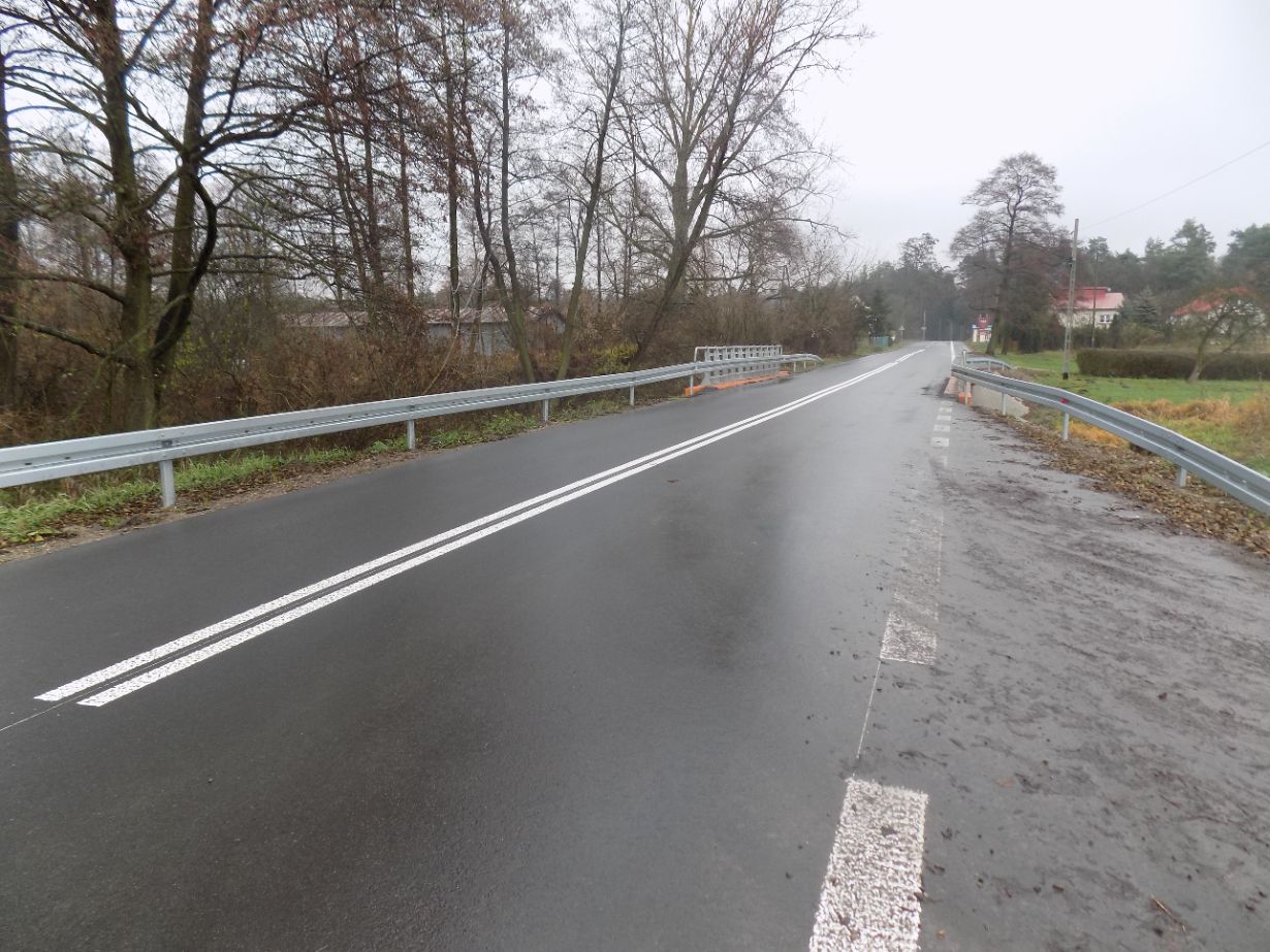 Zdjęcie przedstawia asfaltową drogę z podwójną linią ciągłą pośrodku, prowadzącą przez mostek. Po obu stronach drogi znajdują się metalowe barierki ochronne. W tle widać drzewa i kilka budynków.