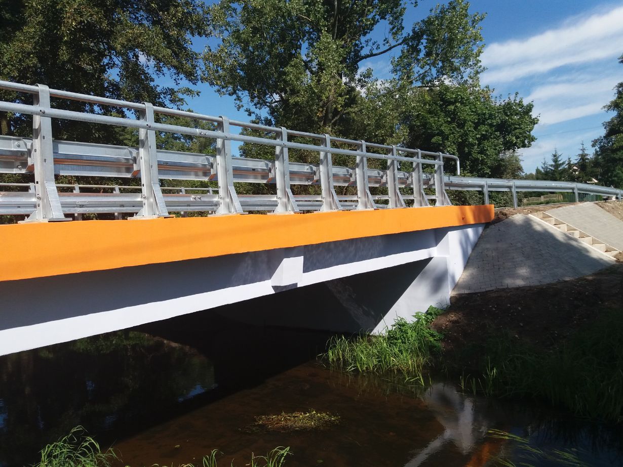 Zdjęcie przedstawia most na rzece z metalowymi barierkami i pomarańczowym pasem na górze. W tle widoczne są drzewa i niebo z chmurami. Po prawej stronie mostu znajdują się schody prowadzące na most.