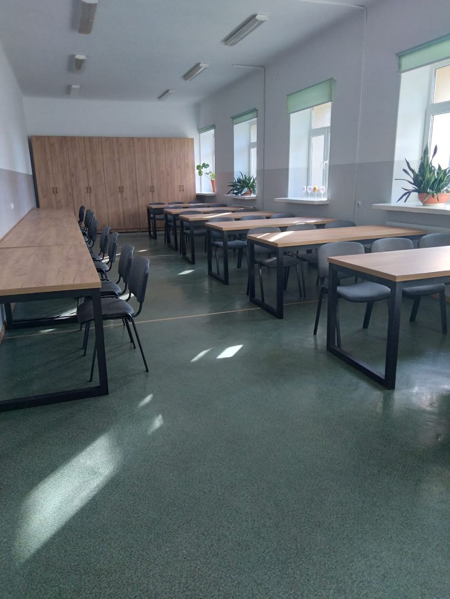 Zdjęcie przedstawia pustą salę lekcyjną z rzędem stołów i krzeseł ustawionych wzdłuż ściany po lewej stronie oraz kilkoma stołami i krzesłami ustawionymi w parach po prawej stronie. Na końcu sali znajdują się drewniane szafy, a na parapetach okien stoją rośliny doniczkowe.