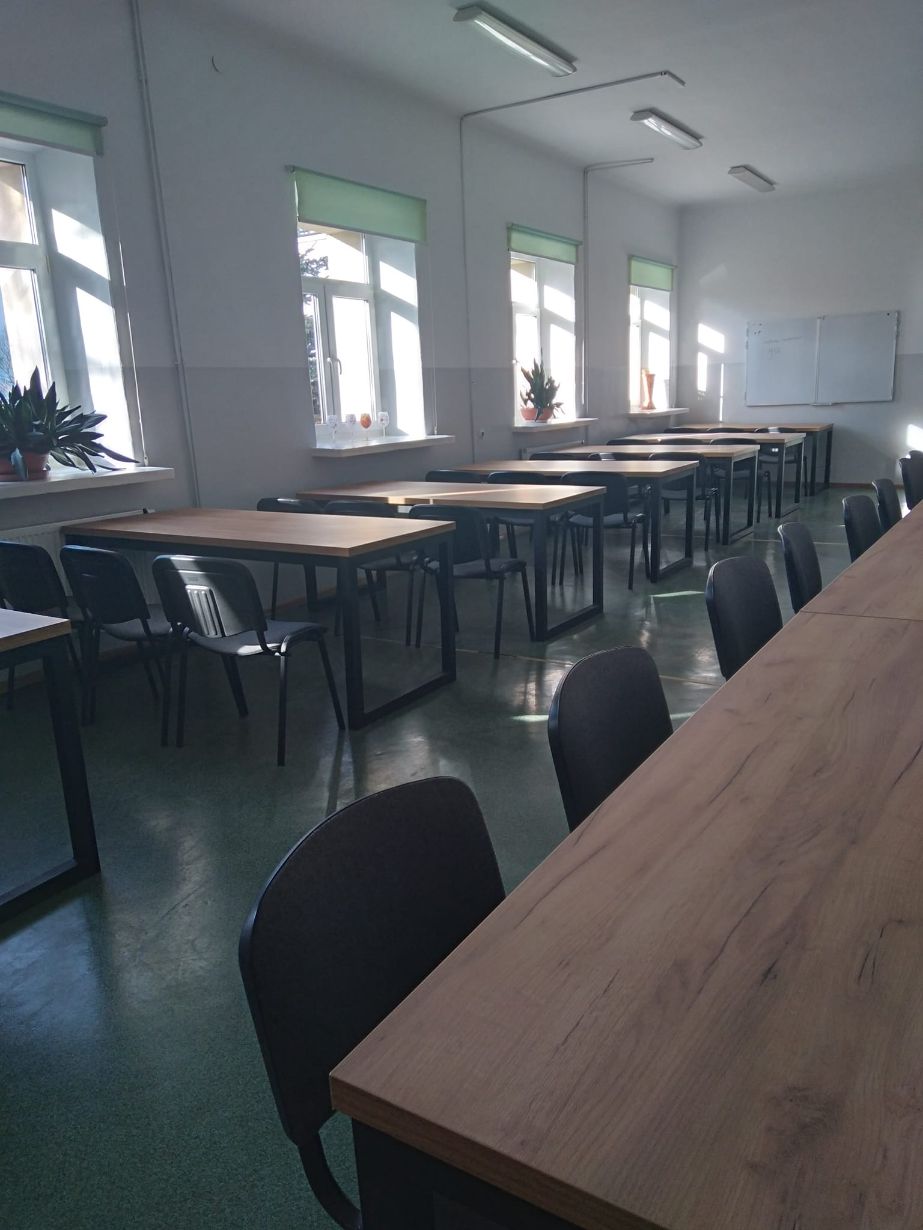 Zdjęcie przedstawia pustą salę lekcyjną z rzędem stołów i krzeseł ustawionych w dwóch rzędach. W sali znajduje się sześć dużych okien z zielonymi roletami, przez które wpada światło dzienne. Na parapetach stoją rośliny doniczkowe. Na ścianie po prawej stronie wisi biała tablica.