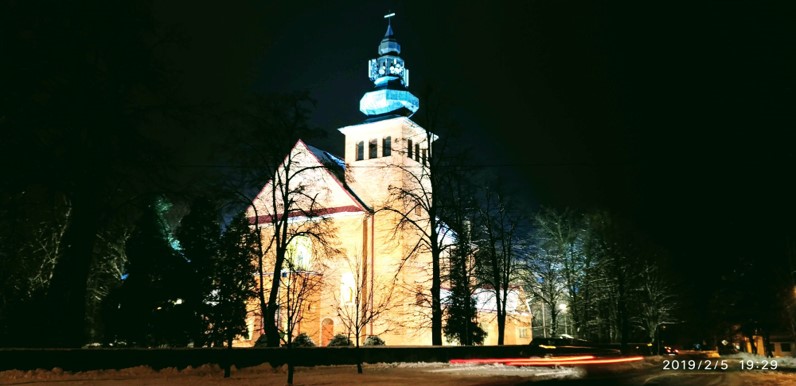 Na zdjęciu widać jasno oświetlony kościół nocą. Kościół ma wysoki szczyt z zegarem i krzyżem na szczycie. Budynek jest oświetlony ciepłymi światłami, podkreślającymi jego architektoniczne cechy. Na pierwszym planie są drzewa, z których niektóre są bez liści, co może wskazywać na zimę. Na ziemi widać śnieg. Niebo jest ciemne, a na drodze przed kościołem widać smugi światła od przejeżdżających pojazdów.