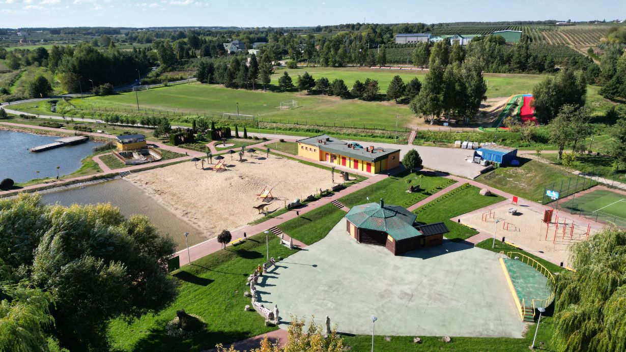 Zdjęcie przedstawia plac zabaw z piaskownicą, huśtawkami i zjeżdżalniami, otoczony zielonymi terenami rekreacyjnymi. W tle widać boisko do piłki nożnej, staw z pomostem oraz budynki gospodarcze.