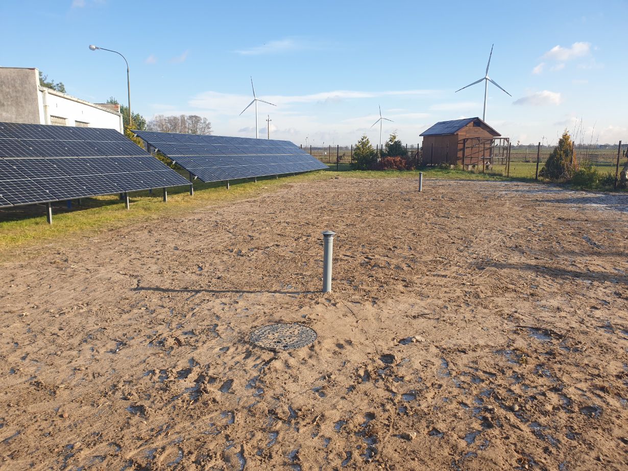 Obraz przedstawia farmę fotowoltaiczną z panelami słonecznymi ustawionymi na trawiastym terenie. W tle widać budynek oraz turbiny wiatrowe. Niebo jest jasne i bezchmurne, co sugeruje słoneczny dzień, idealny do generowania energii słonecznej.