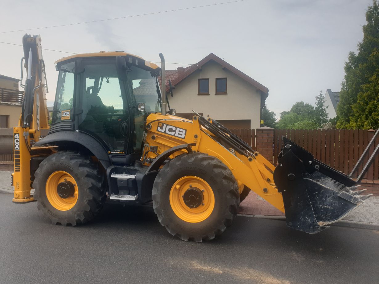 Zdjęcie przedstawia żółto-czarną koparko-ładowarkę marki JCB model 4CX zaparkowaną na ulicy przed domem. W tle widać domy i ogrodzenie.