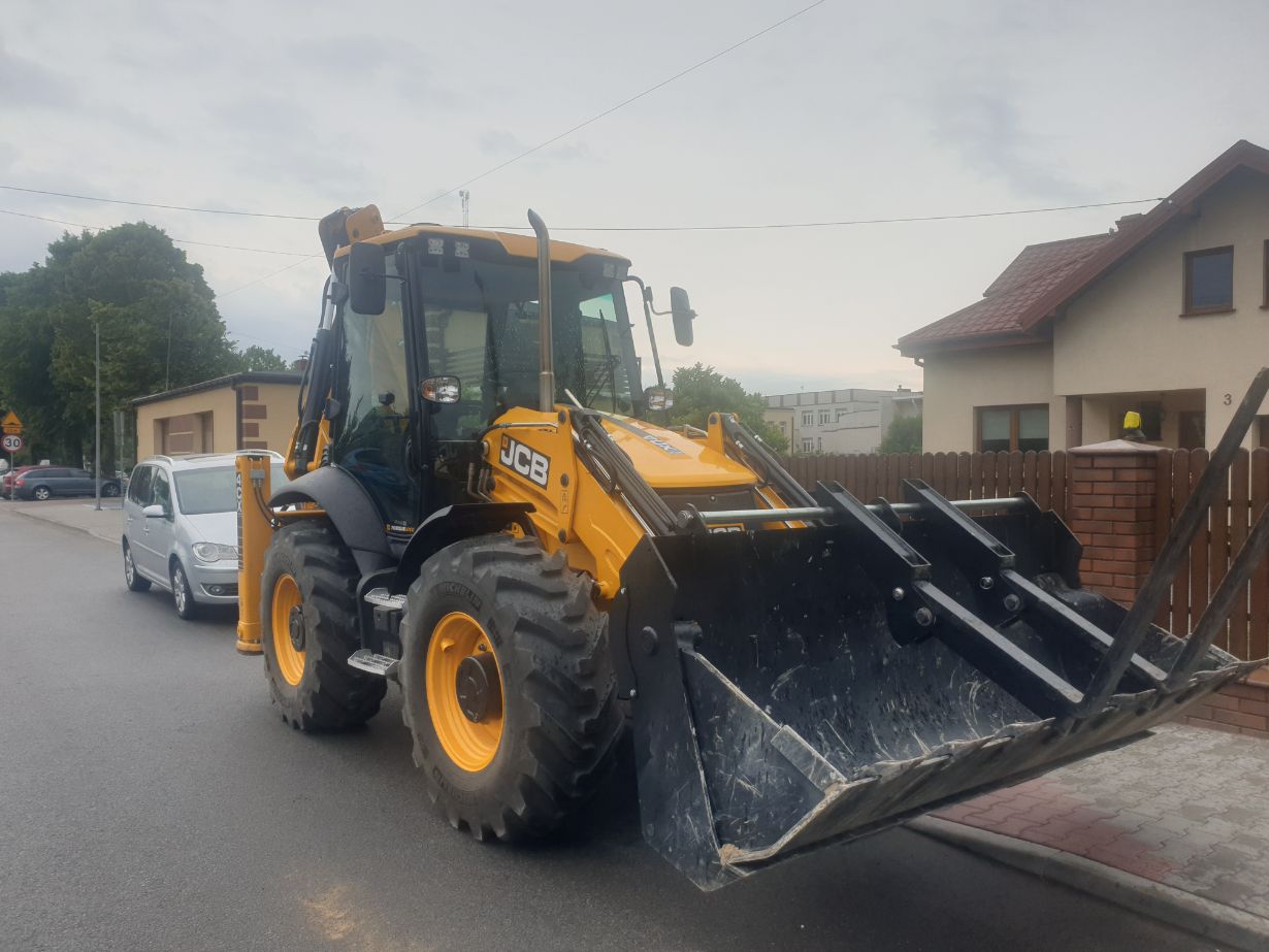 Zdjęcie przedstawia żółtą koparko-ładowarkę marki JCB zaparkowaną na ulicy obok srebrnego samochodu osobowego. W tle widać domy jednorodzinne oraz drzewa.