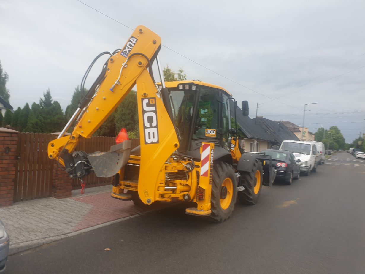 Zdjęcie przedstawia żółtą koparko-ładowarkę marki JCB zaparkowaną na ulicy w pobliżu chodnika. Maszyna ma widoczne ramię z łyżką. W tle znajdują się zaparkowane samochody oraz domy.