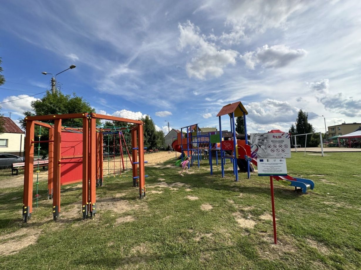 Zdjęcie przedstawia plac zabaw na świeżym powietrzu z różnymi kolorowymi strukturami do zabawy, takimi jak drabinki, zjeżdżalnie i huśtawki. Na pierwszym planie znajduje się tablica z regulaminem placu zabaw. W tle widać budynki mieszkalne i boisko sportowe.