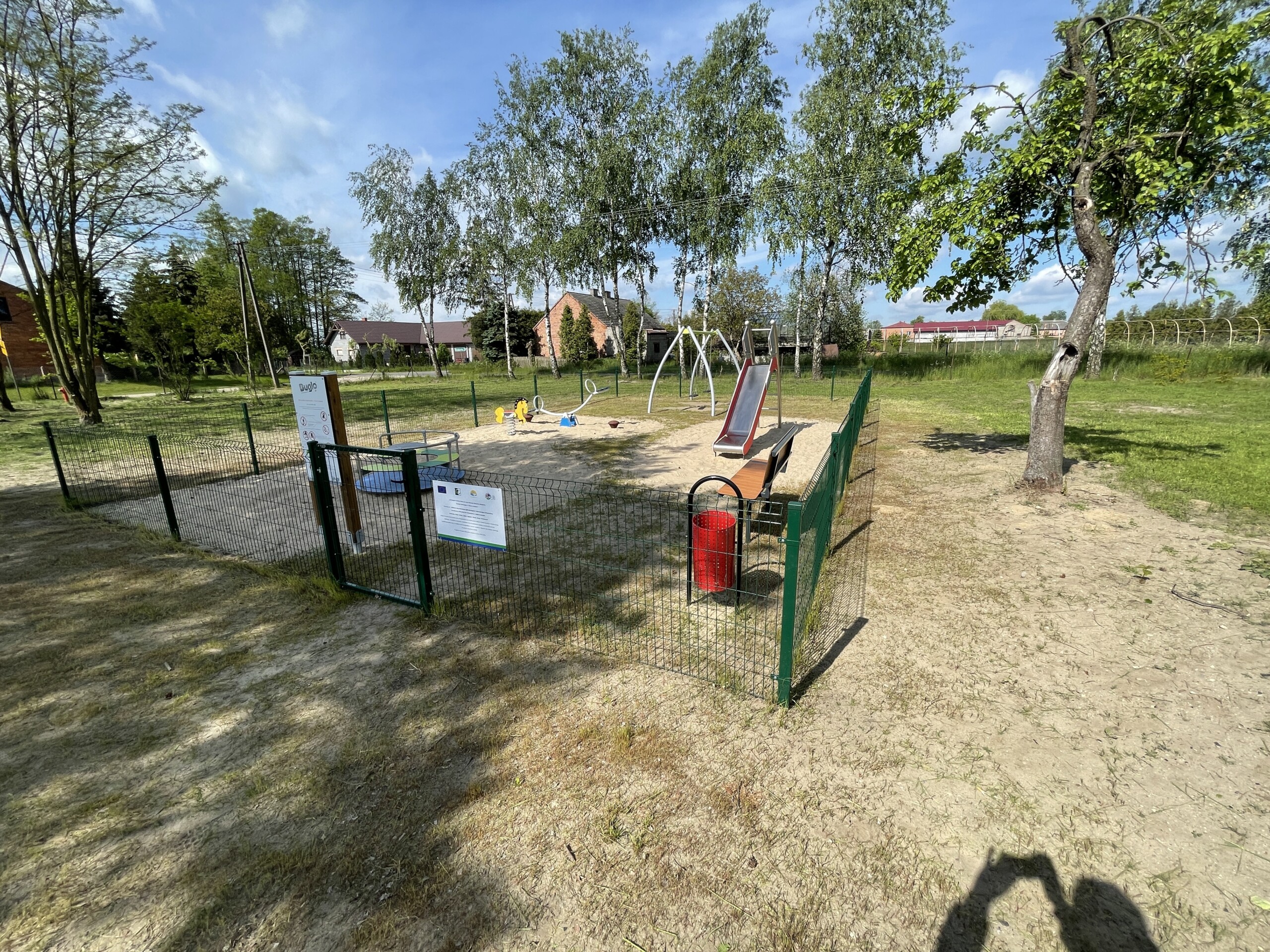 Zdjęcie przedstawia ogrodzony plac zabaw na świeżym powietrzu z piaskownicą, zjeżdżalnią, huśtawkami i innymi urządzeniami do zabawy. W tle widać drzewa, trawę i budynki mieszkalne.