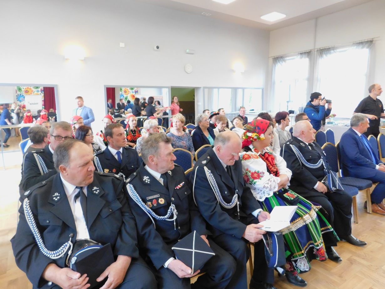 Zdjęcie przedstawia grupę ludzi siedzących w sali, prawdopodobnie podczas jakiegoś wydarzenia lub ceremonii. W pierwszym rzędzie siedzą osoby w mundurach, a obok nich kobieta w tradycyjnym stroju ludowym. W tle widać innych uczestników oraz dekoracje na ścianach.
