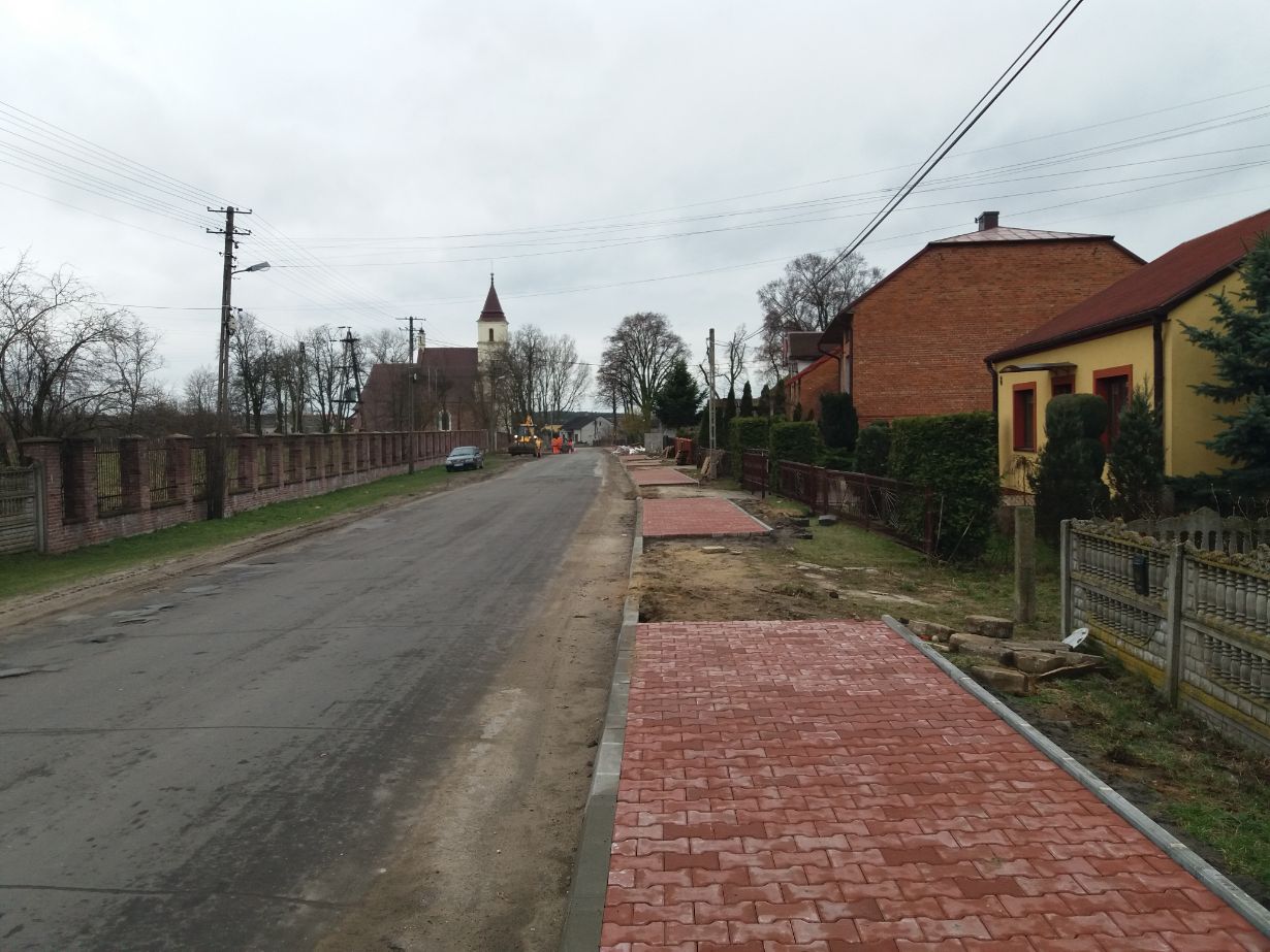 Zdjęcie przedstawia wiejską ulicę z chodnikiem w trakcie budowy. Po lewej stronie znajduje się ogrodzenie z cegły, a po prawej domy z ogrodami. W tle widoczny jest kościół z wieżą.