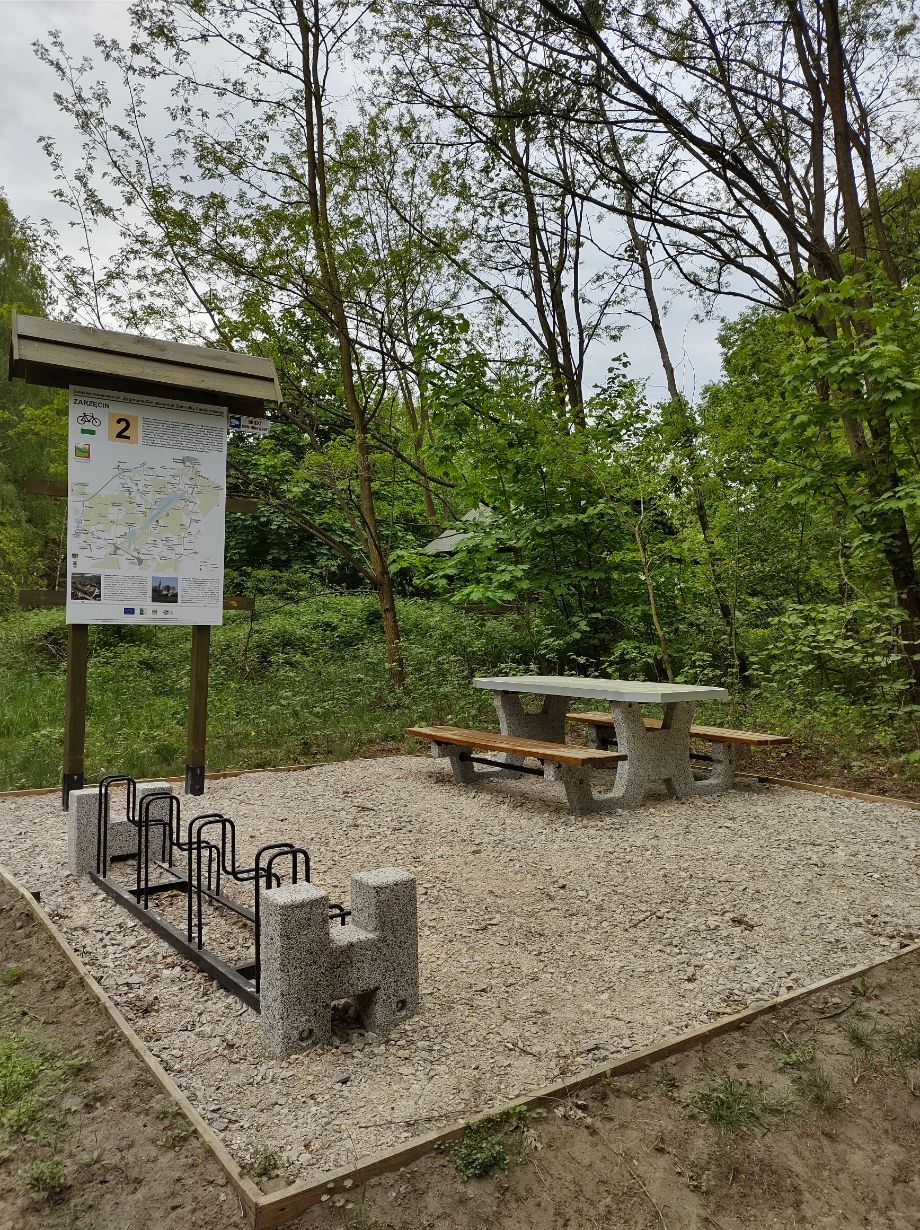 Zdjęcie przedstawia miejsce odpoczynku w lesie z ławką i stołem piknikowym oraz stojakiem na rowery. Obok znajduje się tablica informacyjna z mapą i opisem trasy rowerowej "ZARZĘCIN" oznaczonej numerem 2.