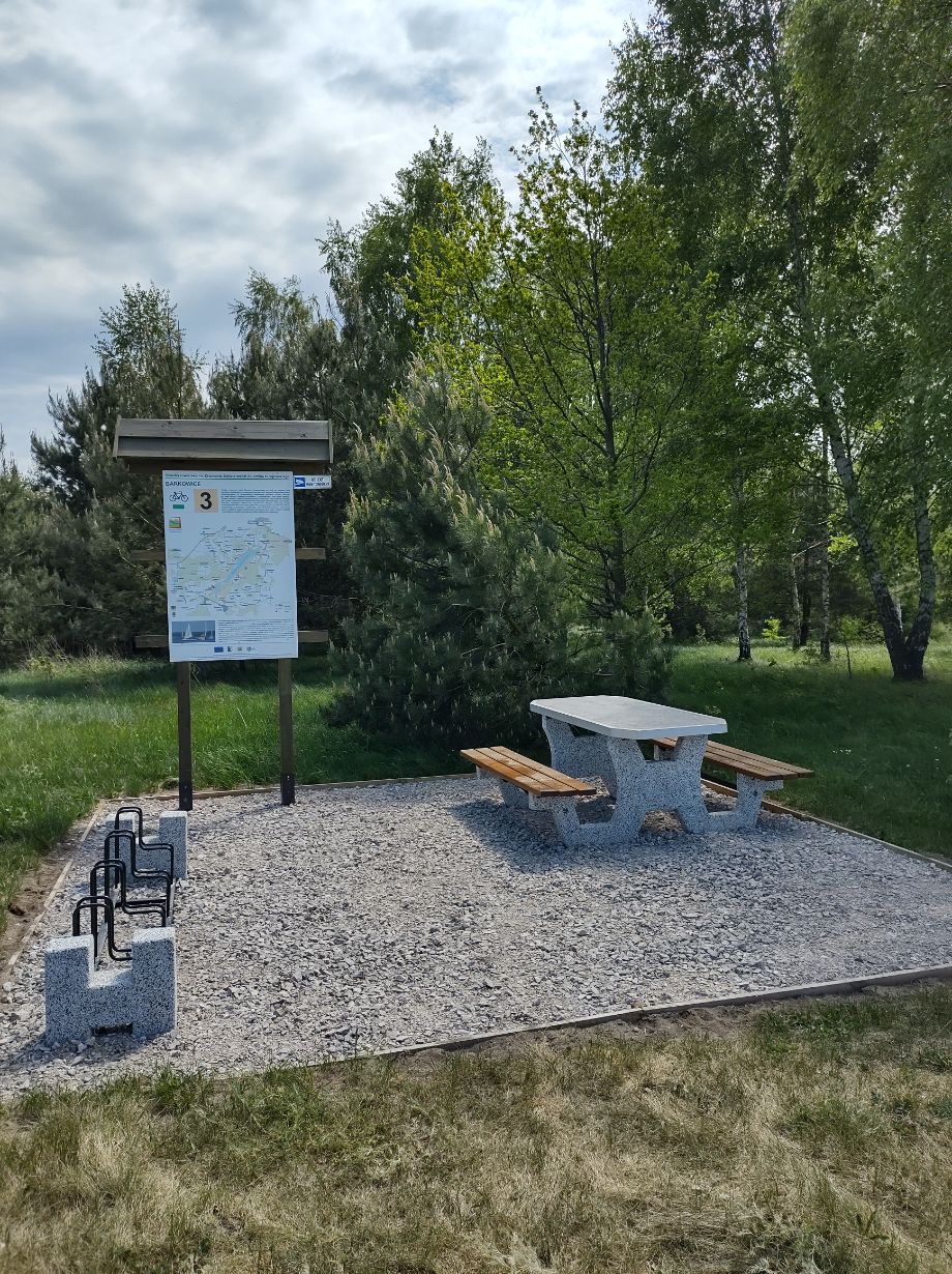 Zdjęcie przedstawia miejsce odpoczynku na szlaku rowerowym. Widać tablicę informacyjną z mapą, stojaki na rowery oraz betonowy stół z ławkami. Otoczenie to zieleń drzew i trawa.