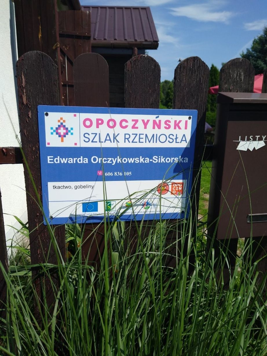 Na zdjęciu znajduje się tabliczka informacyjna na drewnianym płocie. Tabliczka informuje o "Opoczyńskim Szlaku Rzemiosła" i zawiera dane kontaktowe Edwardy Orczykowskiej-Sikorskiej, zajmującej się tkactwem i gobelinami.