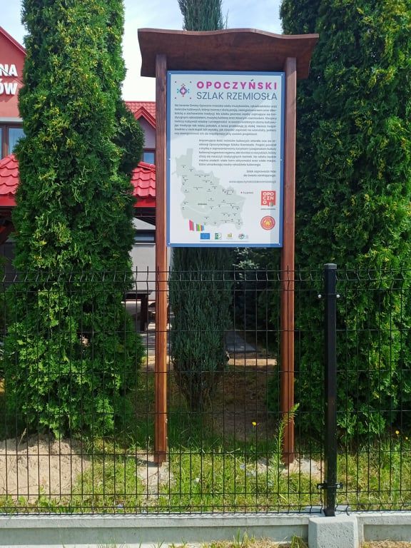 Na zdjęciu widać tablicę informacyjną "Opoczyński Szlak Rzemiosła" umieszczoną na drewnianej konstrukcji, otoczoną zielonymi drzewami i ogrodzeniem. Na tablicy znajduje się mapa oraz tekst opisujący szlak rzemiosła w regionie Opoczna.