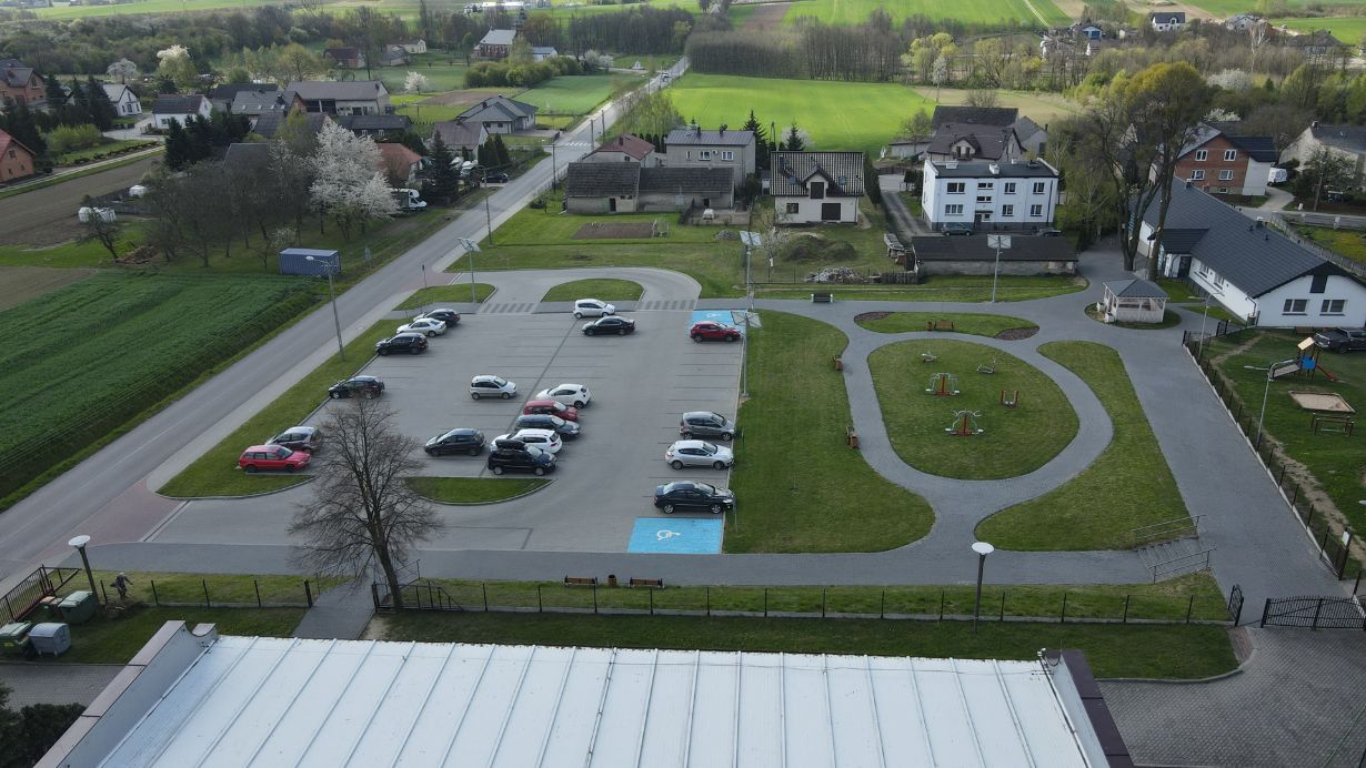 Zdjęcie przedstawia parking z kilkunastoma samochodami, w tym dwa miejsca dla osób niepełnosprawnych oznaczone niebieskim kolorem. Obok parkingu znajduje się plac zabaw z trawiastymi ścieżkami i kilkoma urządzeniami do zabawy. W tle widać domy mieszkalne, pola uprawne i zalesione tereny.