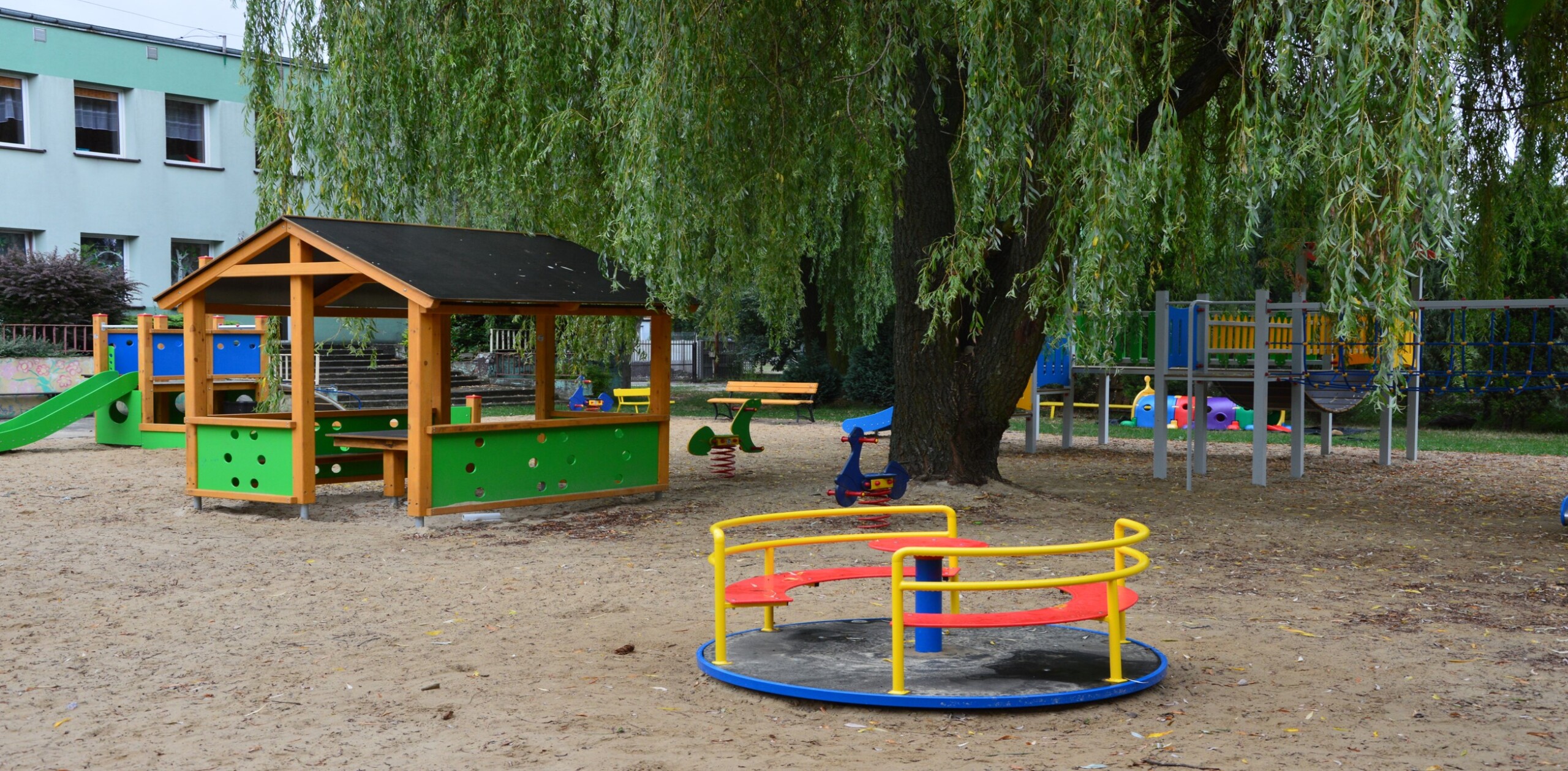 Na zdjęciu znajduje się plac zabaw z różnymi elementami do zabawy. Widać drewnianą altankę z zielonymi panelami, karuzelę z żółtymi poręczami i czerwonymi siedzeniami, zjeżdżalnię, huśtawki na sprężynach oraz drabinki. W tle rośnie duże drzewo z opadającymi gałęziami.