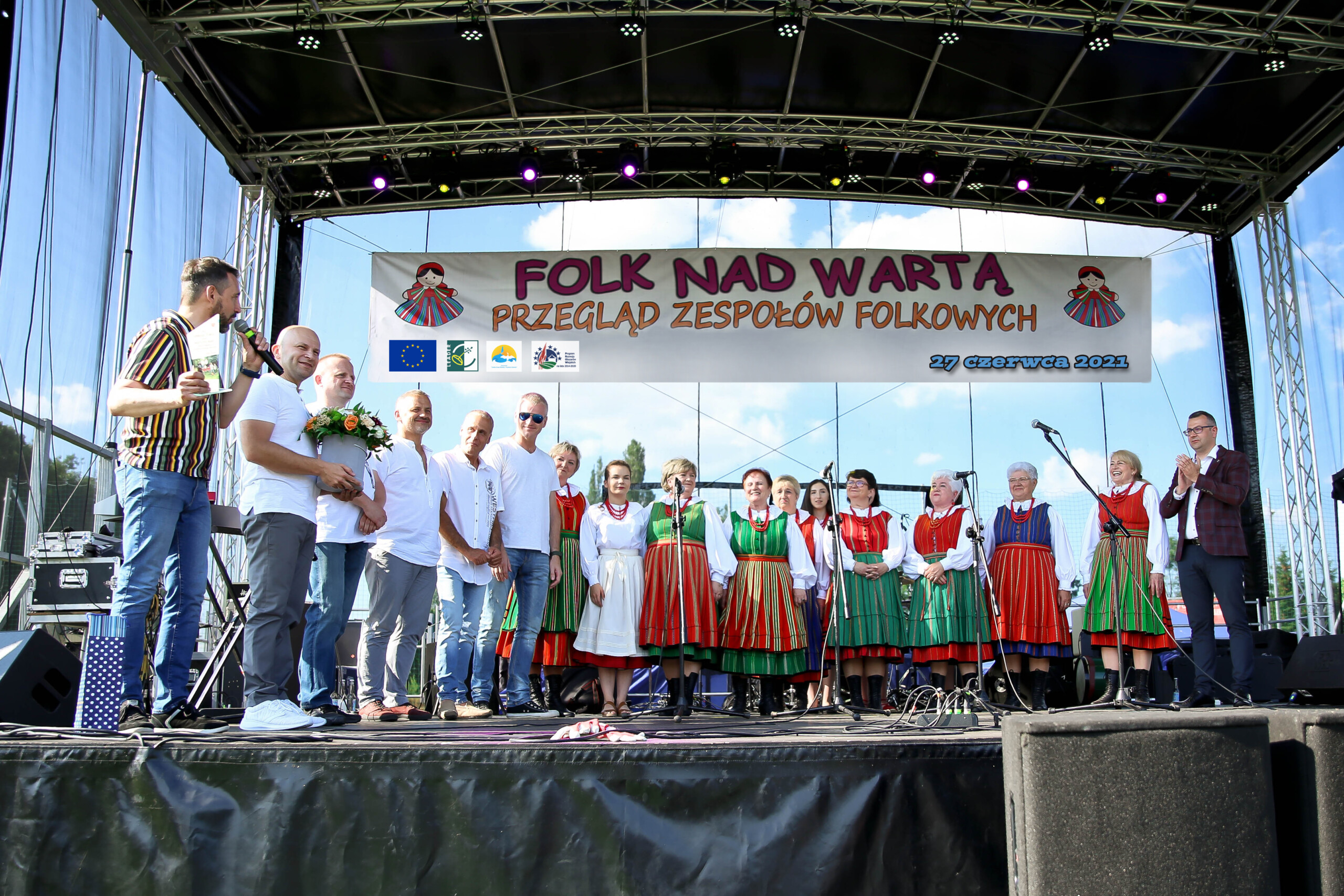 Na scenie znajduje się grupa ludzi ubranych w tradycyjne stroje ludowe, uczestniczących w wydarzeniu "Folk nad Wartą - Przegląd Zespołów Folkowych" z dnia 27 czerwca 2021 roku. Nad sceną wisi baner z nazwą wydarzenia oraz informującymi o finansowaniu projektu z UE.