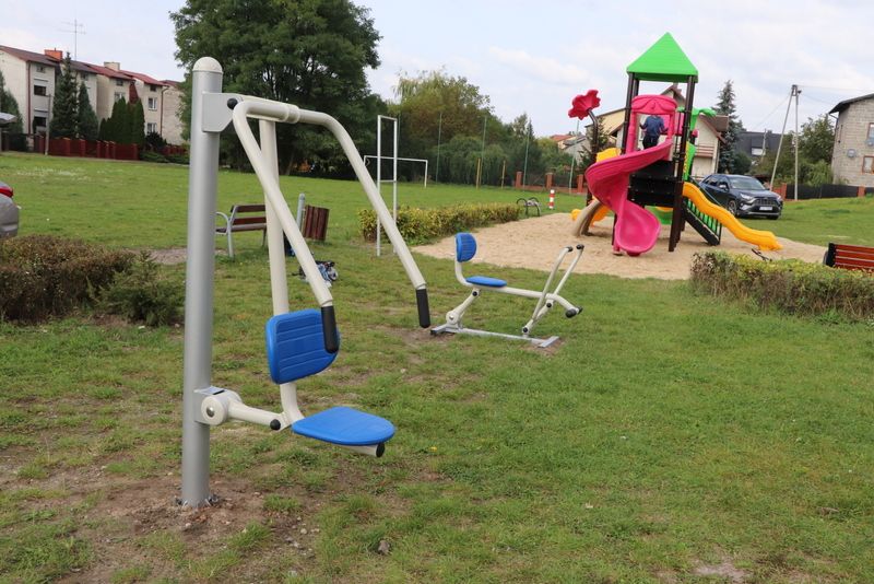 Na zdjęciu znajduje się plac zabaw na świeżym powietrzu z różnymi urządzeniami do ćwiczeń i zabawy. Na pierwszym planie widoczne są dwa urządzenia do ćwiczeń siłowych z niebieskimi siedzeniami. W tle znajduje się kolorowa zjeżdżalnia z wieżą, na której bawią się dzieci.