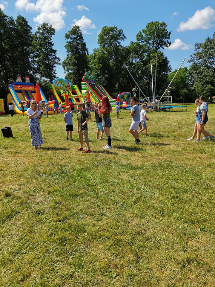 Grupa ludzi, w tym dzieci i dorośli, zgromadzona na zewnątrz przy dmuchanych zjeżdżalniach i trampolinie bungee. Scena przypomina wesołe miasteczko lub wydarzenie społecznościowe w słoneczny dzień, z zieloną trawą i drzewami w tle.