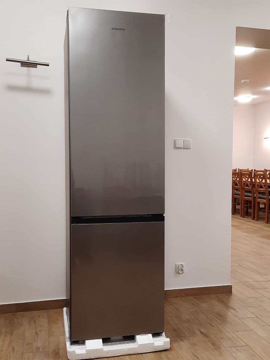 Na zdjęciu widać drewnianą altanę z zadaszeniem, stojącą na wyłożonym kostką brukową placu. Obok altany znajduje się okrągłe palenisko z cegieł. W tle widoczne są budynki i trawniki.