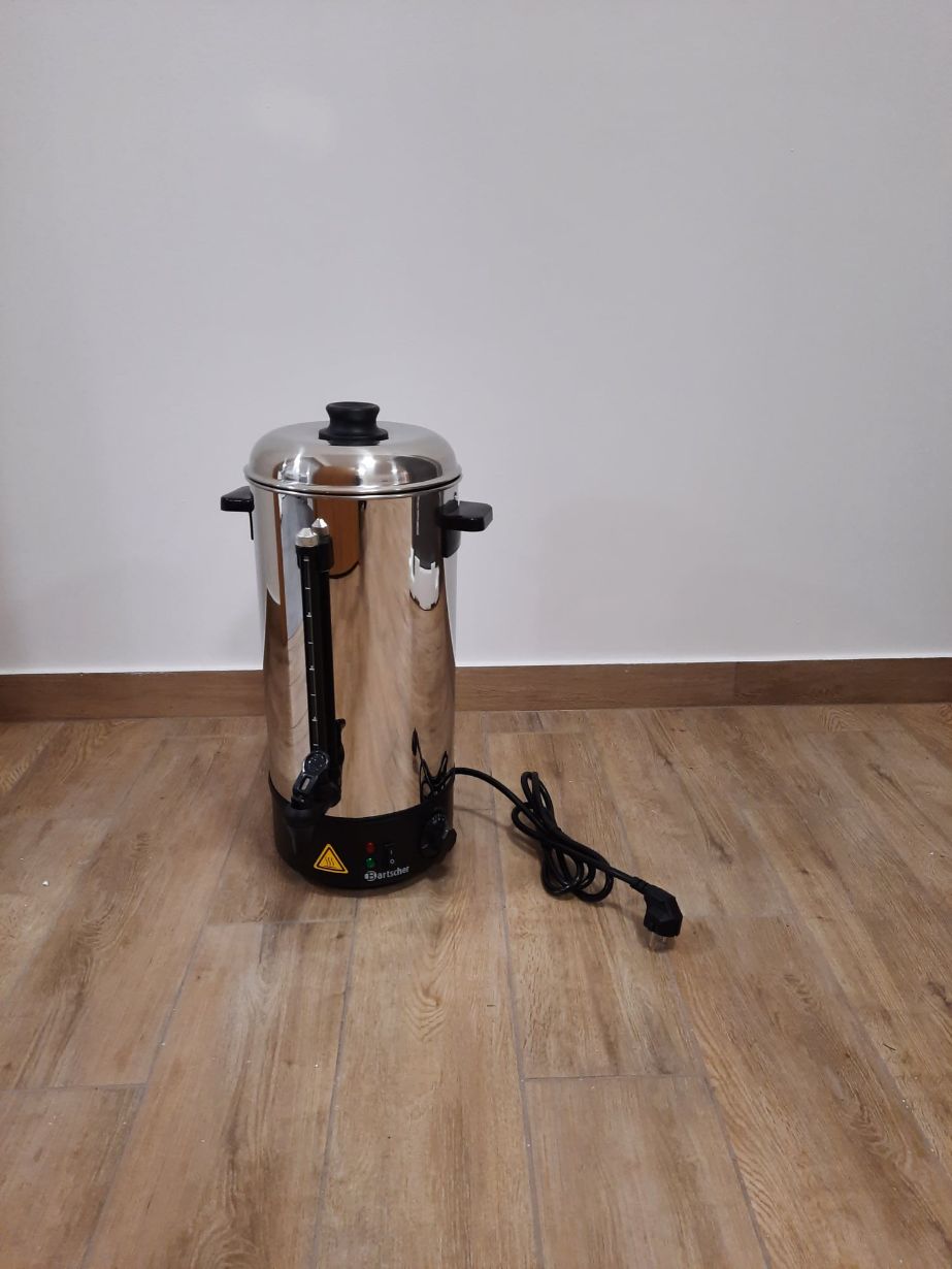 Na zdjęciu widać stalowy elektryczny czajnik z czarną pokrywką i uchwytami, ustawiony na drewnianej podłodze przy białej ścianie. Czajnik ma wskaźnik poziomu wody i podłączony przewód zasilający.
