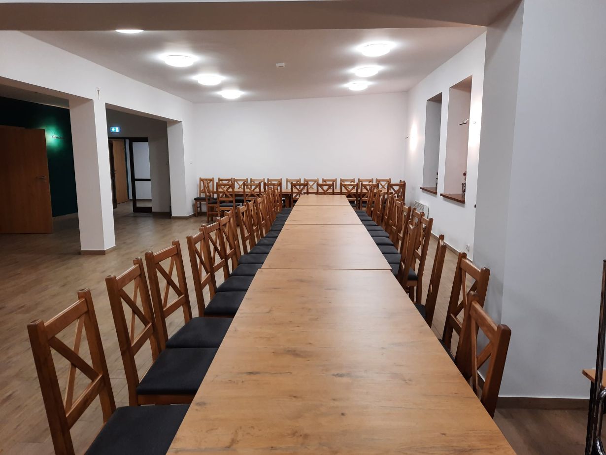 Na zdjęciu widać długi drewniany stół otoczony wieloma drewnianymi krzesłami z czarnymi poduszkami. W tle widać otwarte drzwi prowadzące do innego pomieszczenia oraz kilka okien na ścianie po prawej stronie.