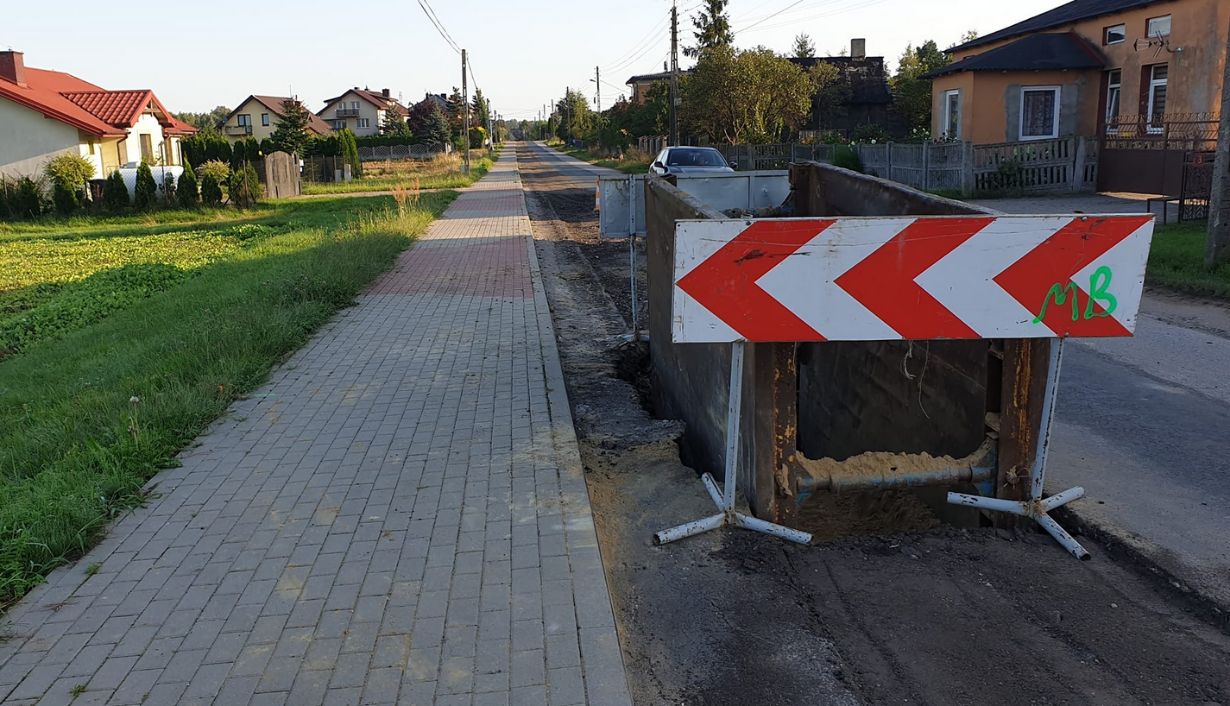 Zdjęcie przedstawia ulicę w małej miejscowości, na której trwają prace drogowe. Po prawej stronie znajduje się wykop zabezpieczony metalowymi barierami, na których umieszczono znak ostrzegawczy z czerwonymi strzałkami skierowanymi w lewo. Po lewej stronie widać chodnik z kostki brukowej oraz trawnik. W tle znajdują się domy jednorodzinne, drzewa i zaparkowany samochód. Zdjęcie jest interesujące, ponieważ pokazuje typowy widok prac drogowych w małej miejscowości, co może być istotne dla mieszkańców i użytkowników drogi.