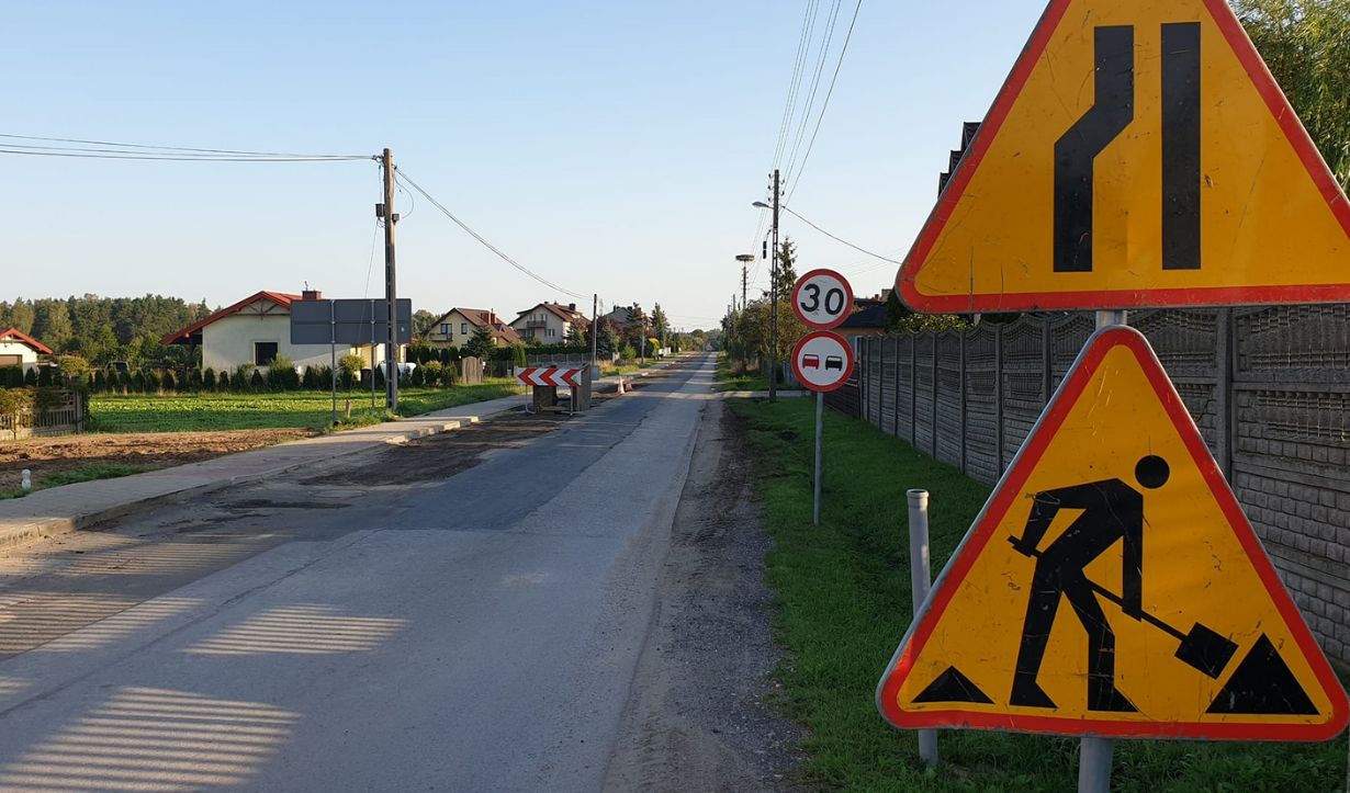 Zdjęcie przedstawia wiejską drogę z kilkoma znakami drogowymi. Na pierwszym planie po prawej stronie znajdują się dwa trójkątne znaki ostrzegawcze: jeden informujący o zwężeniu jezdni, a drugi o robotach drogowych. Obok nich znajduje się okrągły znak ograniczenia prędkości do 30 km/h oraz zakaz wyprzedzania. W tle widać domy jednorodzinne, pola uprawne oraz słupy energetyczne. Droga jest częściowo rozkopana, co sugeruje trwające prace remontowe.