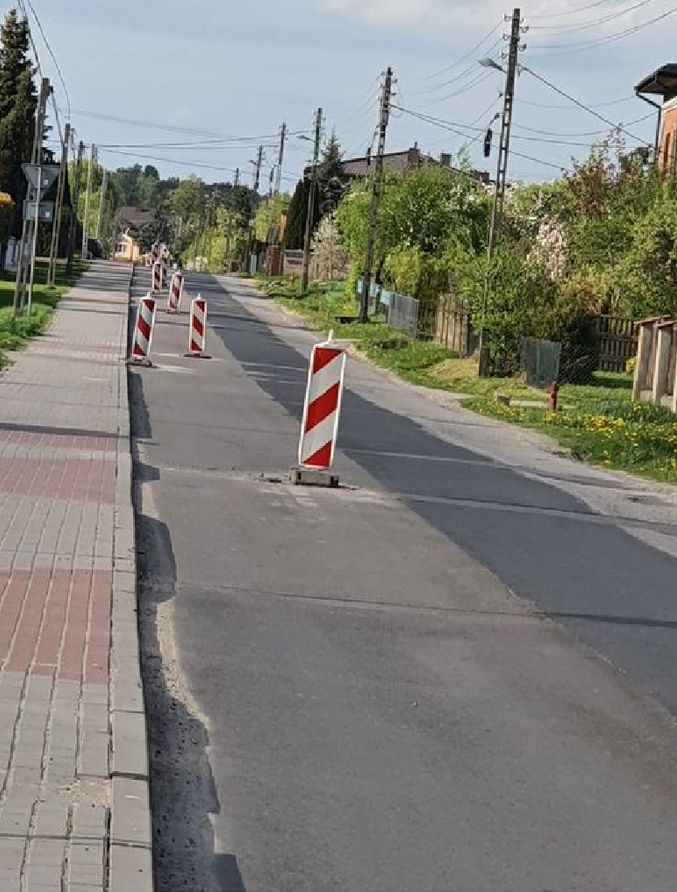 Zdjęcie przedstawia ulicę w trakcie remontu. Na jezdni ustawione są biało-czerwone słupki ostrzegawcze, które wskazują na prace drogowe lub naprawy nawierzchni. W tle widać domy, drzewa oraz słupy energetyczne z przewodami. Po lewej stronie znajduje się chodnik z kostki brukowej.