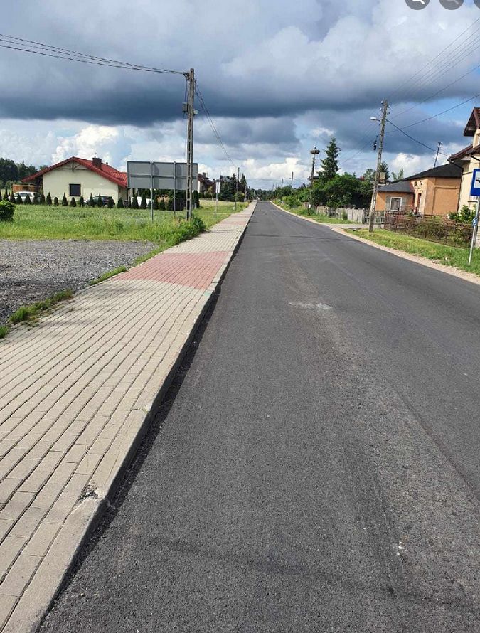 Zdjęcie przedstawia prostą, asfaltową drogę biegnącą w dal, z chodnikiem po lewej stronie. Chodnik jest wykonany z kostki brukowej i ma fragmenty w kolorze czerwonym. Po obu stronach drogi znajdują się domy jednorodzinne z ogrodami. Na niebie widać ciemne, burzowe chmury. W tle, po lewej stronie, znajduje się znak drogowy. Droga jest pusta, bez pojazdów ani pieszych.