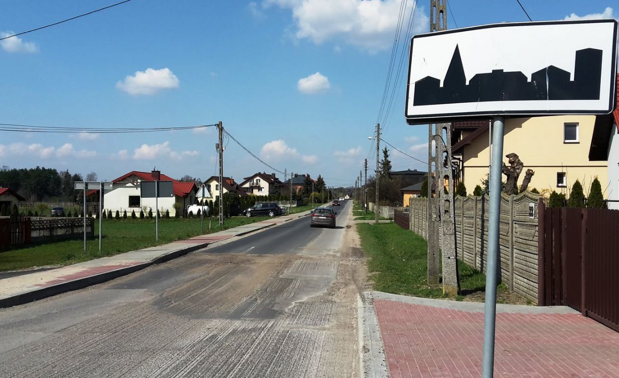 Zdjęcie przedstawia wiejską drogę biegnącą przez małą miejscowość. Po obu stronach drogi znajdują się domy jednorodzinne z ogrodami. Na pierwszym planie po prawej stronie widoczny jest znak drogowy z sylwetką miasta, co sugeruje, że wjeżdżamy do obszaru zabudowanego. Droga jest asfaltowa, ale widać na niej ślady naprawy lub remontu. Po lewej stronie drogi znajduje się chodnik z czerwonej kostki brukowej. Na drodze widoczne są dwa samochody jadące w kierunku przeciwnym do obserwatora. W tle widać drzewa i niebo z kilkoma chmurami.