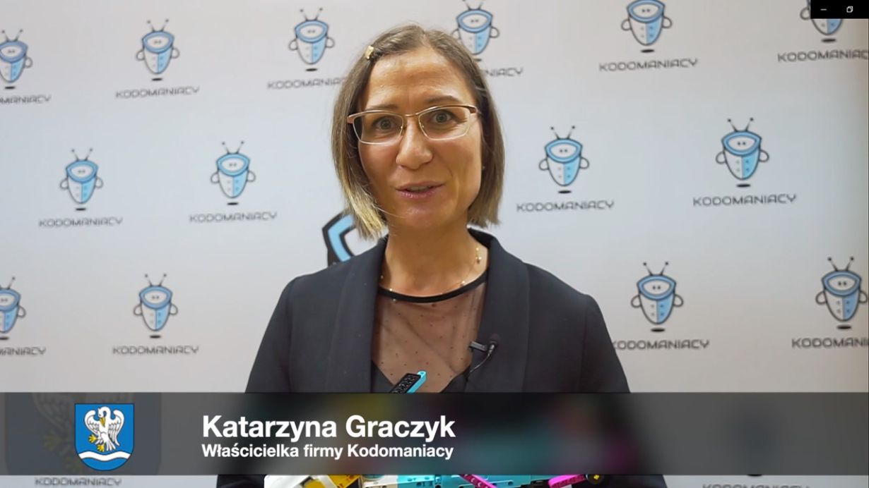 Na zdjęciu znajduje się kobieta na tle ścianki z logo Kodomaniacy. Na dole ekranu widnieje pasek z napisem: "Katarzyna Graczyk Właścicielka firmy Kodomaniacy". Obok napisu znajduje się herb z białym orłem na niebieskim tle.
